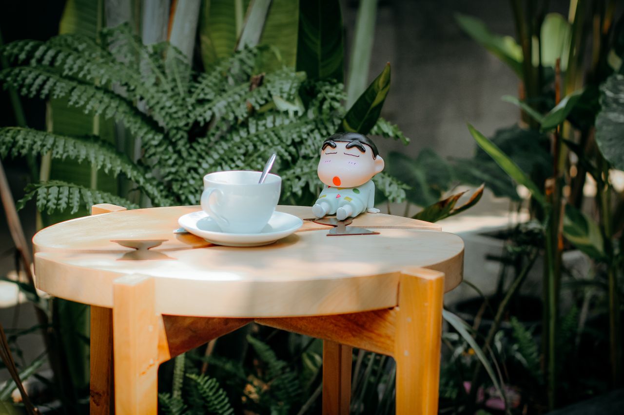 hero-img A serene outdoor setting with a coffee cup and cartoon figurine on a wooden table surrounded by greenery.